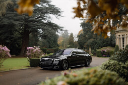 Black luxury Mercedes-Benz sedan parked in a gated estate driveway with manicured gardens and a stately building in the background