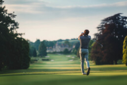 Golfer in striped shirt swinging a club on a green golf course with trees in the background.