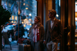 Couple with rolling suitcases stands outside a shop at dusk, smiling and looking to the side in smart coats and scarfed outfits.