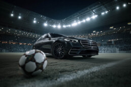 Black Mercedes-Benz sedan on a soccer field inside a stadium at night, with bright floodlights overhead and a blurred soccer ball in the foreground.