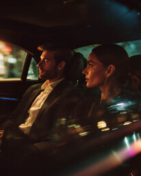 A man and a woman sit in a dim car at night, city lights blurring outside the windows as they ride in calm silence.