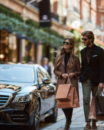 Stylish couple wearing sunglasses walks on a city sidewalk carrying shopping bags beside a black luxury car.