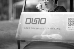 Driver wearing glasses sits in a chauffeured vehicle with a banner reading 'YOUR CHAUFFEUR, ON DEMAND' on the side.