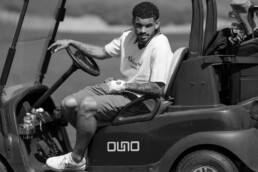 Man seated in a golf cart on a golf course, looking toward the camera with tattoos on his forearm and wearing a white polo and shorts.