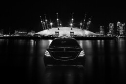 Rear view of a glossy black Mercedes-Benz on a dark night, with a lit domed stadium and city skyline in the background.