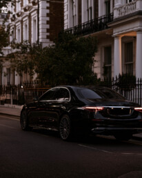 Black luxury sedan parked on a city street in front of white townhouse buildings at dusk.