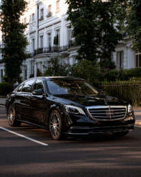 Black Mercedes sedan parked on a tree-lined city street with white row houses in the background.