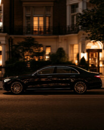 Black luxury sedan travels along a city street at dusk, illuminated brownstone facades glowing in the windows.