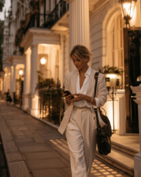 Woman in a light beige suit checks her phone while standing beside a columned building at dusk on a city street.