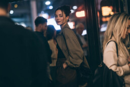 Young woman with a bun and tan trench coat glances back over her shoulder in a dimly lit night-time city scene, standing by glass doors as people mingle behind her.