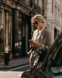 Woman in a beige suit and sunglasses stands on a city street looking at her phone.