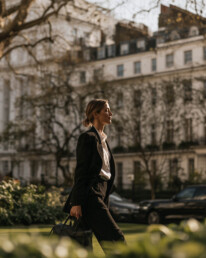 Professional woman in a black blazer walks along a city street with greenery and white apartment buildings in the background.