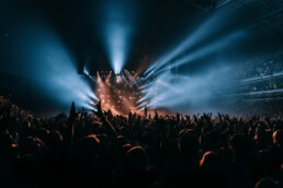 Crowd at a concert with raised hands in front of a brightly lit stage and blue atmospheric lighting.