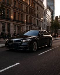 Black luxury sedan driving down a city street at dusk with tall buildings on both sides, headlights on, urban scene behind.
