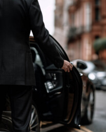 Person in a black suit opens the back door of a dark car on a city street.