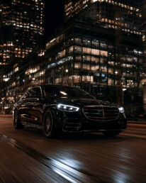 Black luxury sedan speeding through a neon-lit city street at night, with blurred sidewalks and tall glass buildings behind it.