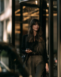Businesswoman in a black blazer and brown trousers stands beside a glass building, looking to the side while holding a smartphone.